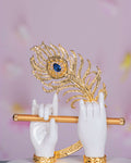 A close-up of Lord Krishna's hands, in white, holding a golden flute and a detailed golden peacock feather adorned with a blue gem.