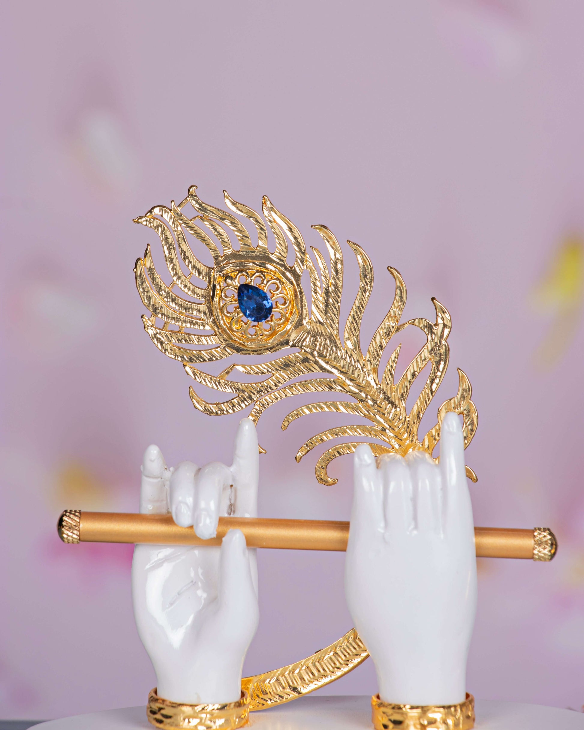 A close-up of Lord Krishna's hands, in white, holding a golden flute and a detailed golden peacock feather adorned with a blue gem.