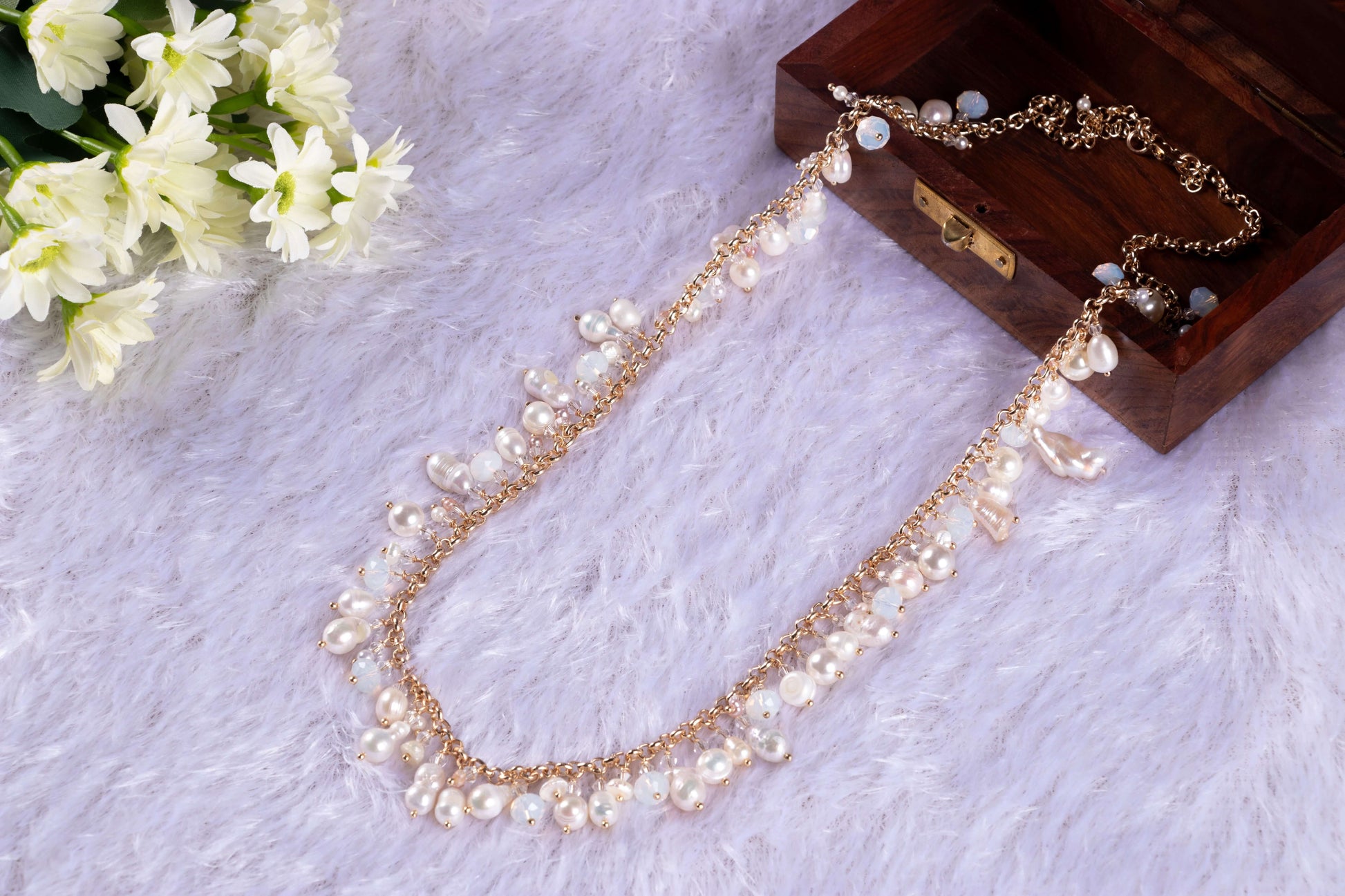 A long gold-toned necklace featuring an array of Baroque and mother-of-pearl (MOP) shell pearls of varying sizes and shapes, displayed on a white furry surface next to a small wooden box and white flowers.