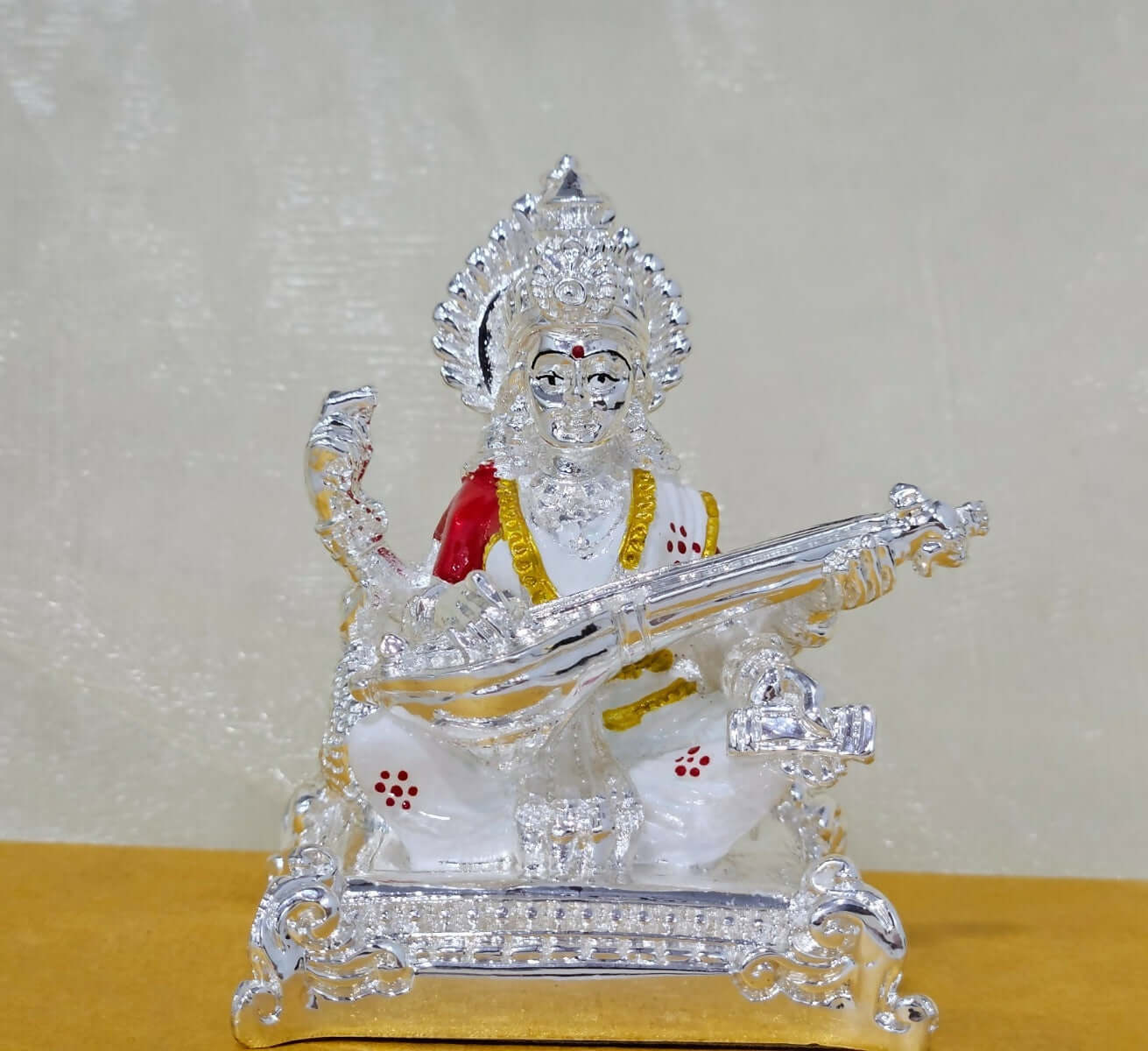 Silver-plated idol of Goddess Saraswati seated and playing the veena, with accents of red and gold on her attire.