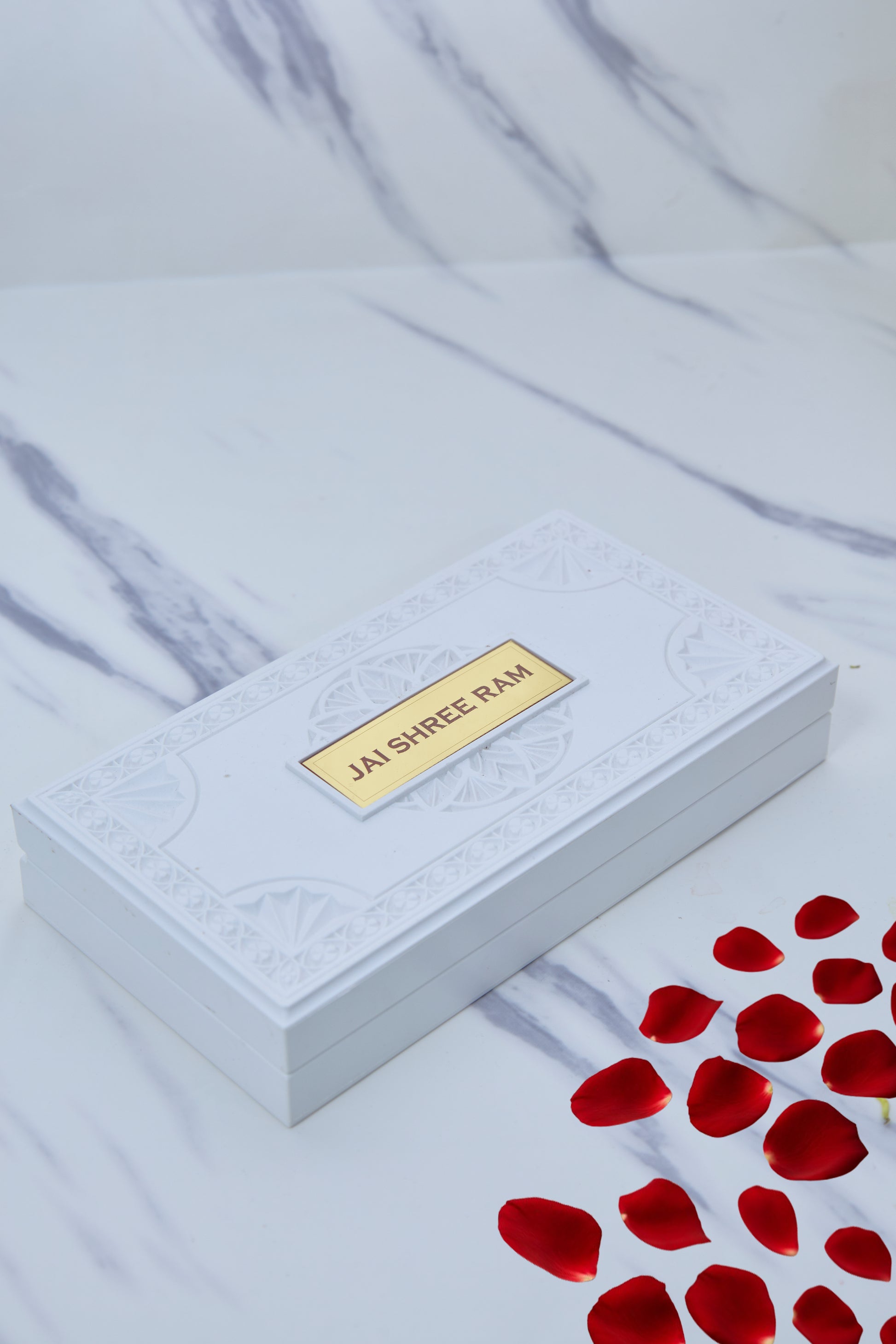 An image featuring a sleek white box, likely a "Ram Darbar Pocket Temple," with a prominent gold-colored plate on its lid that reads "JAI SHREE RAM" in capitalized letters. The box is adorned with subtle, embossed patterns on its lid. Scattered in the foreground and to the right are vibrant red rose petals, adding a touch of devotion and aesthetic appeal. The background is a soft, marbled white surface.