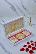 A white open box, revealing a golden depiction of the Ram Darbar (Lord Rama, Sita, Lakshmana, and Hanuman) on the inner lid. Inside the box, a golden Charan Paduka (footprints) and two smaller golden coins with inscriptions rest on a red velvet-lined base. A decorative golden bell with a feather-like top and scattered red rose petals are visible in the background, all placed on a white marble-patterned surface.