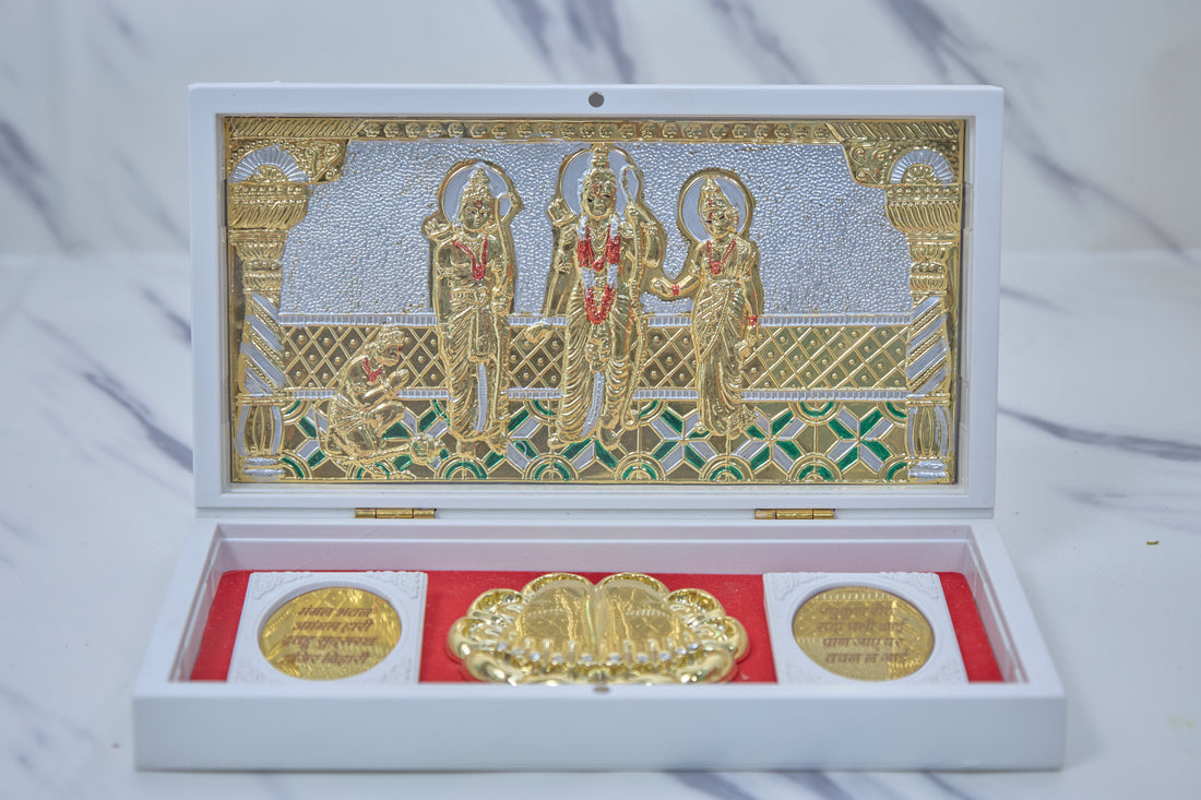 A white box open to reveal a gold-plated Ram Darbar tableau inside, featuring Lord Rama, Sita, Lakshmana, and Hanuman, alongside golden religious coins on a red velvet base.
