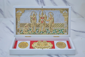 A white box open to reveal a gold-plated Ram Darbar tableau inside, featuring Lord Rama, Sita, Lakshmana, and Hanuman, alongside golden religious coins on a red velvet base.