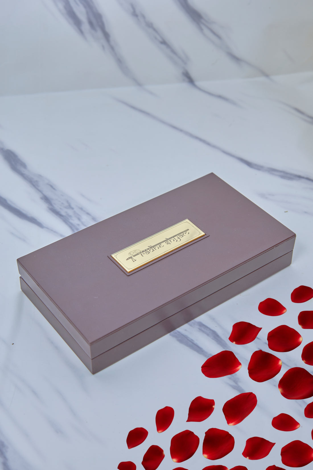 An open wooden gift box with a red velvet interior containing Hindu devotional items, including a gold-plated image of Radha Krishna on the inside lid, a pair of golden Charan Paduka (divine footprints), and two framed plaques with text, alongside a small golden bell outside the box.