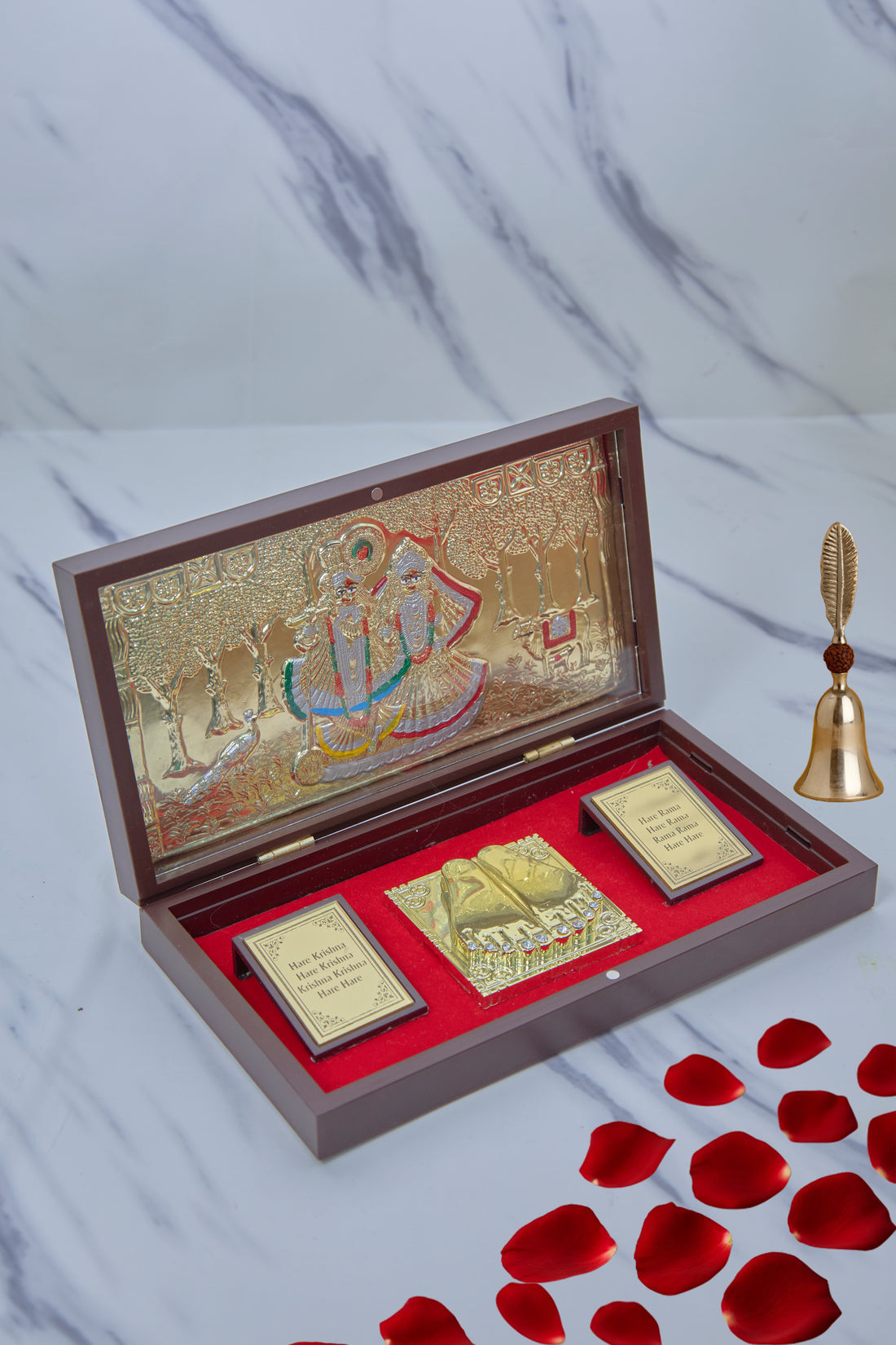 An open wooden gift box with a red velvet interior containing Hindu devotional items, including a gold-plated image of Radha Krishna on the inside lid, a pair of golden Charan Paduka (divine footprints), and two framed plaques with text, alongside a small golden bell outside the box.