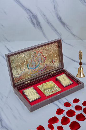 An open wooden gift box with a red velvet interior containing Hindu devotional items, including a gold-plated image of Radha Krishna on the inside lid, a pair of golden Charan Paduka (divine footprints), and two framed plaques with text, alongside a small golden bell outside the box.