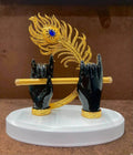 A decorative statue featuring Lord Krishna's dark-skinned hands holding a golden flute, with a vibrant golden peacock feather with a blue eye detail rising behind the hands, all set on a white oval base.