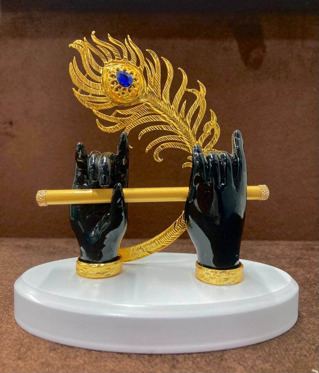 A decorative statue featuring Lord Krishna's dark-skinned hands holding a golden flute, with a vibrant golden peacock feather with a blue eye detail rising behind the hands, all set on a white oval base.