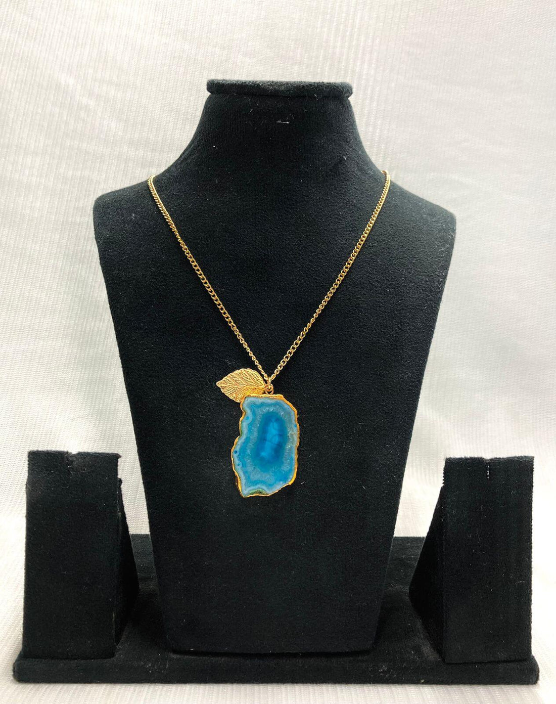 A gold-colored chain necklace featuring a blue agate leaf pendant, displayed on a black stand.