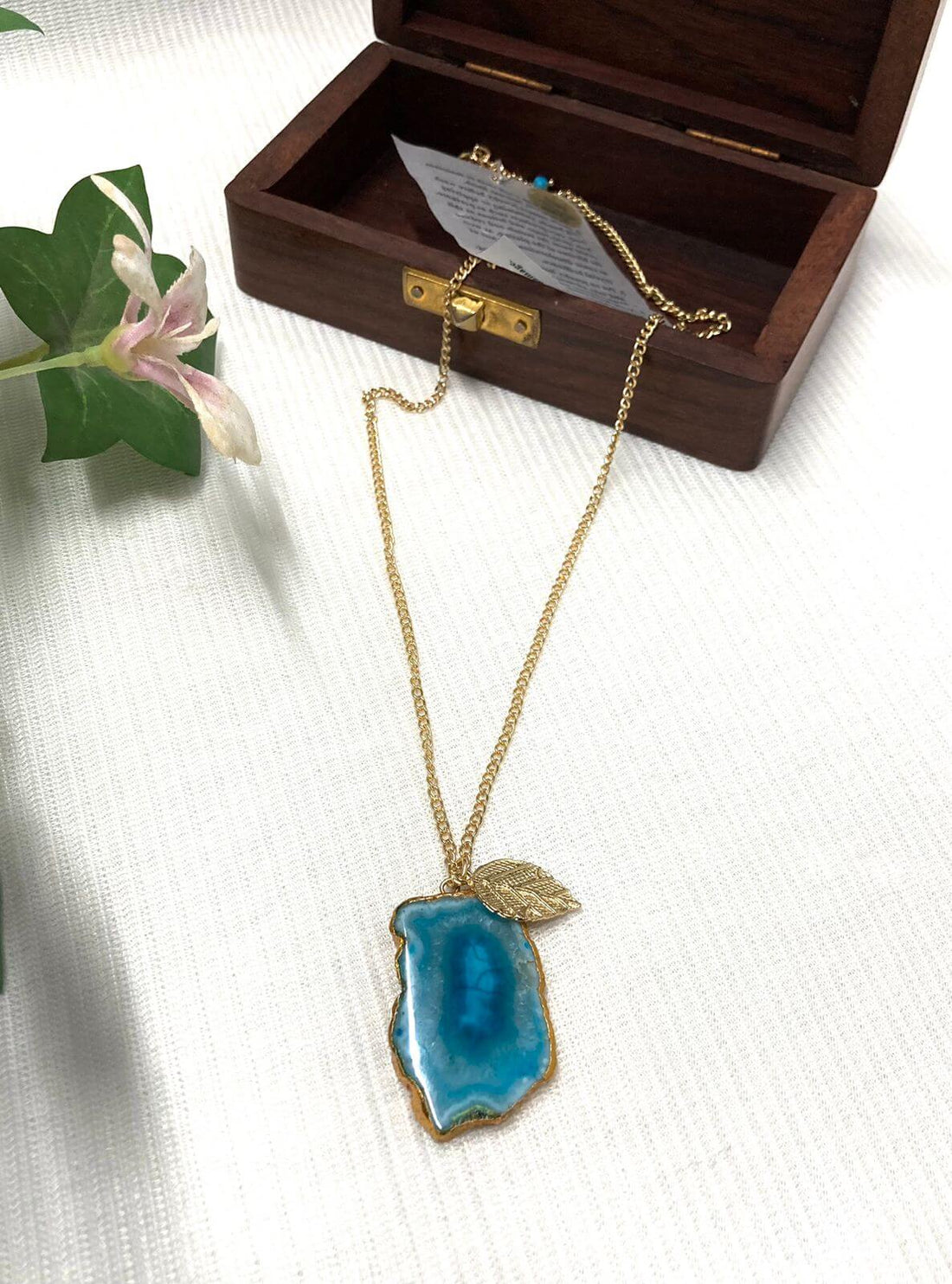 A gold-colored chain necklace featuring a blue agate leaf pendant, displayed on a black stand.