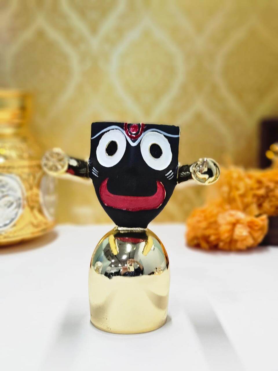 A photograph of a Jagannath Idol, which is 4 inches tall, placed on a golden base with a serene countenance and intricate detailing, against a plain backdrop.