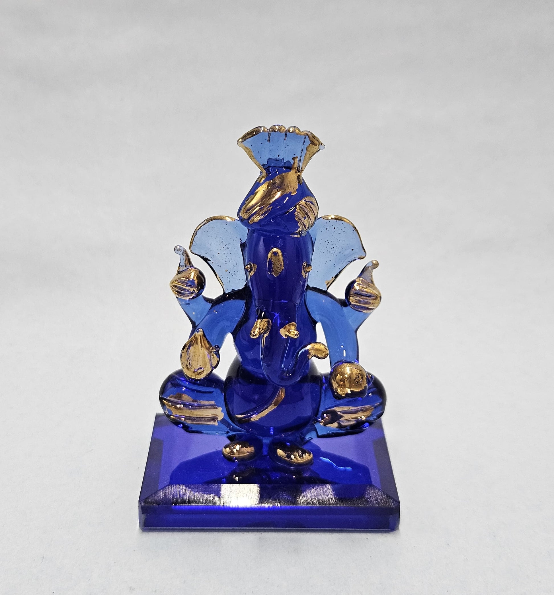 A small, intricately designed blue and gold Pagri Ganesha idol made of glass or crystal, seated on a rectangular blue base.