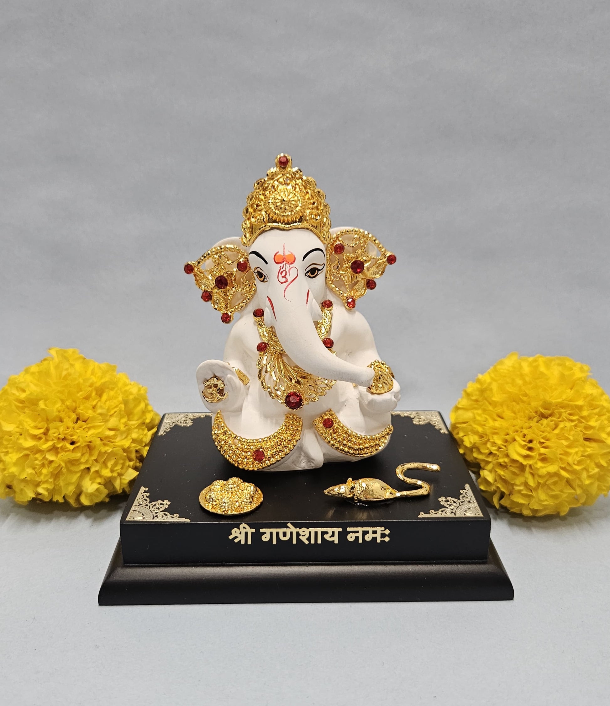 A white and gold Ganesha idol, adorned with red jewels and intricate golden patterns, seated on a black base inscribed with "शà¥à¤°à¥€ गणेशाय नमः" in Hindi script. The base also features small golden accents and a golden mouse figurine. Two bright yellow marigold flower arrangements flank the idol on either side, all set against a neutral grey background.