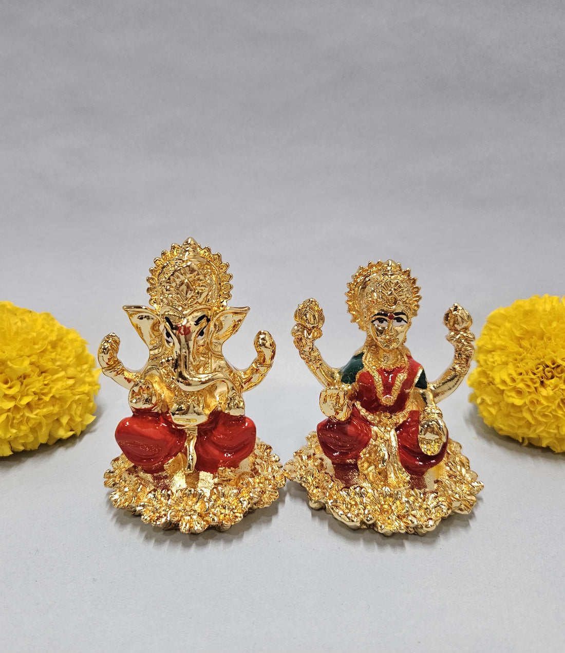 Gold-plated idols of Lord Ganesha and Goddess Lakshmi, seated on lotus-like bases, flanked by vibrant yellow marigold flowers on a light background.