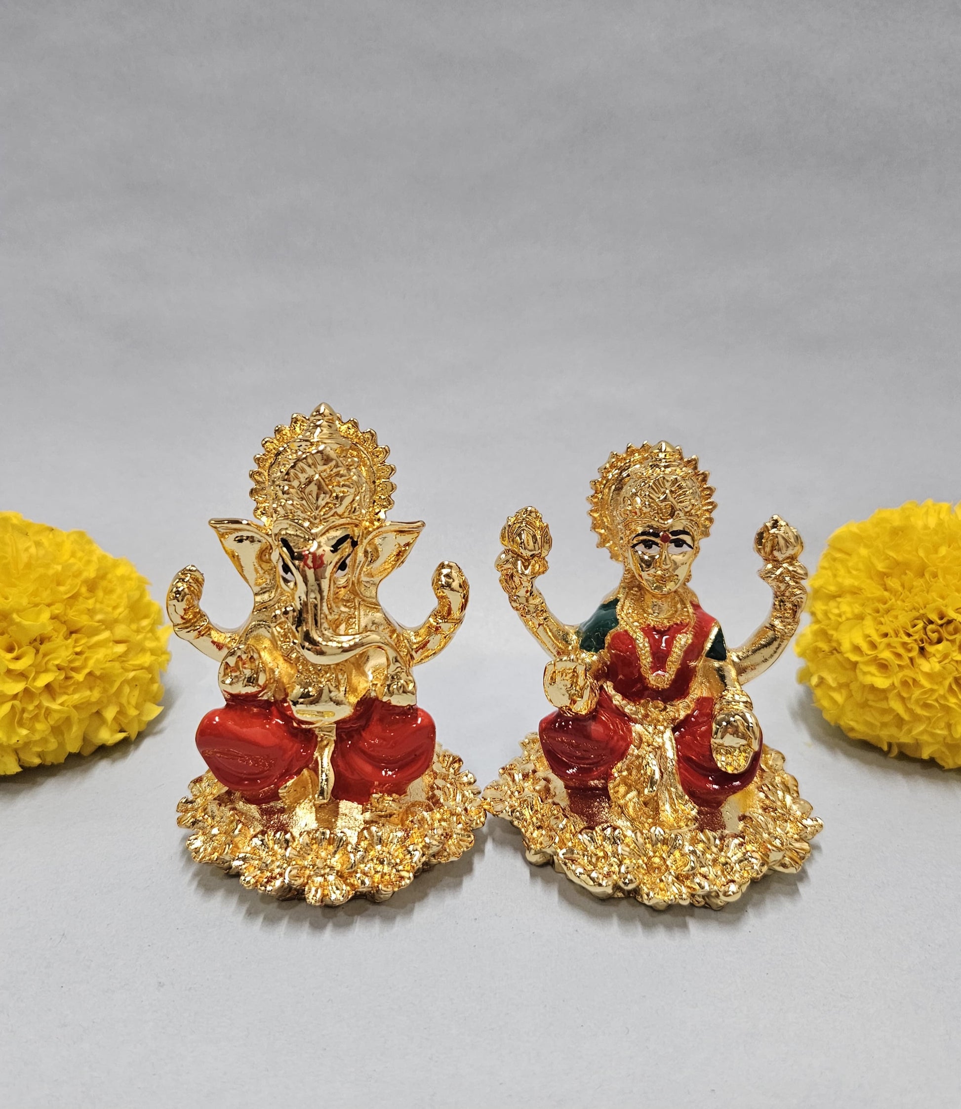 Gold-plated idols of Lord Ganesha and Goddess Lakshmi, seated on lotus-like bases, flanked by vibrant yellow marigold flowers on a light background.