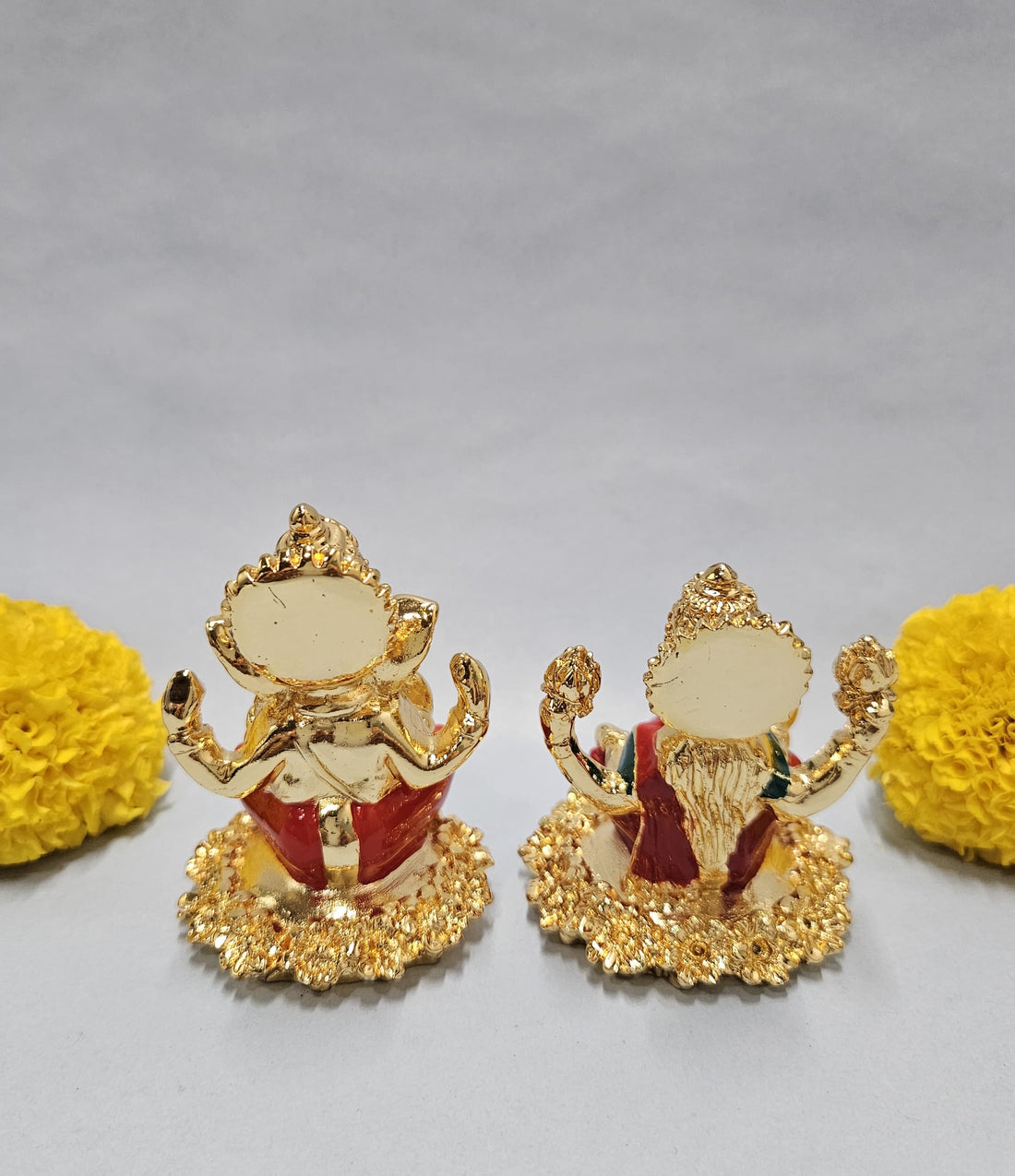 Gold-plated idols of Lord Ganesha and Goddess Lakshmi, seated on lotus-like bases, flanked by vibrant yellow marigold flowers on a light background.
