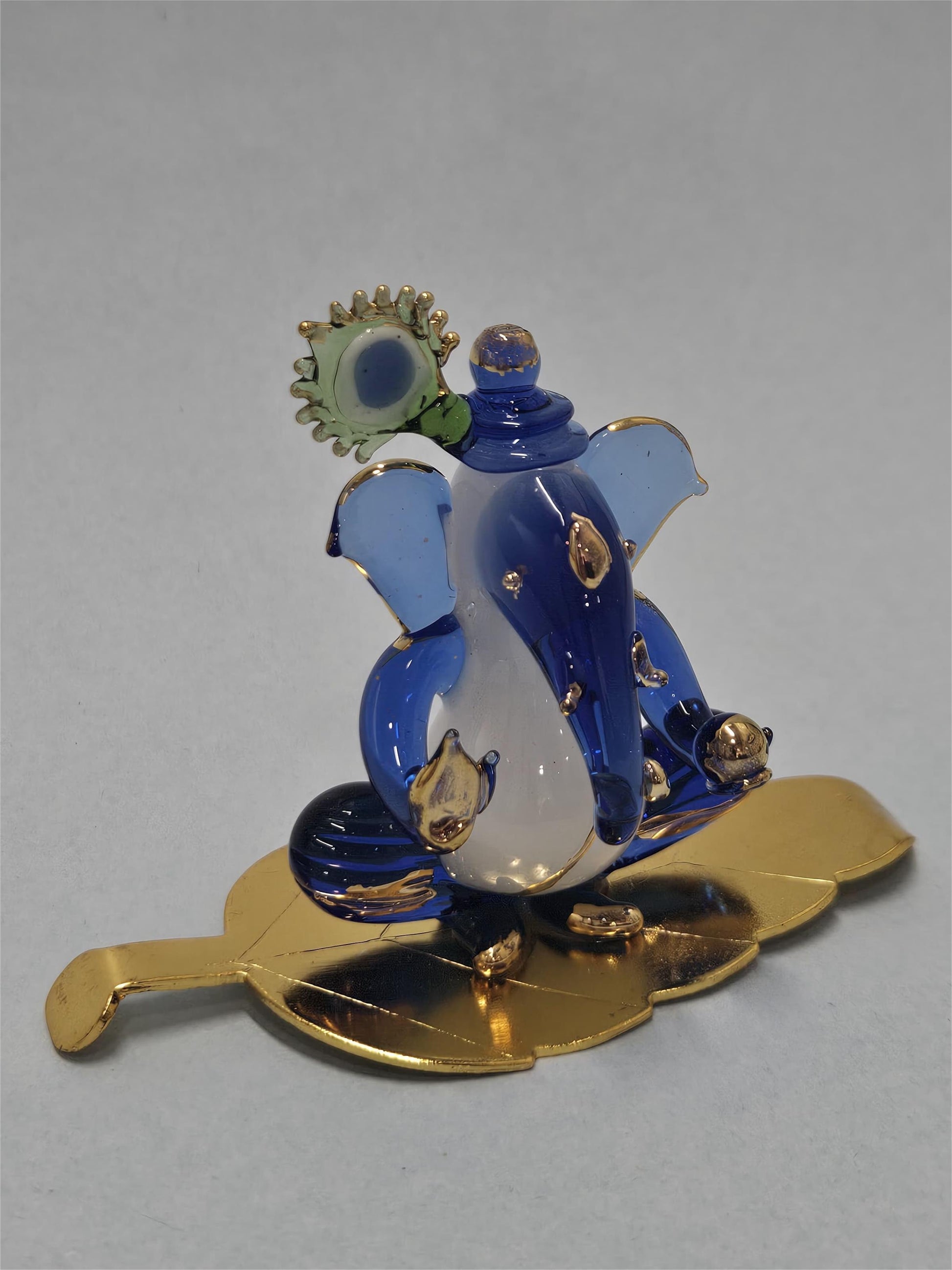 A blue and white glass Morpankhi Ganesha idol sitting on a gold-polished leaf.