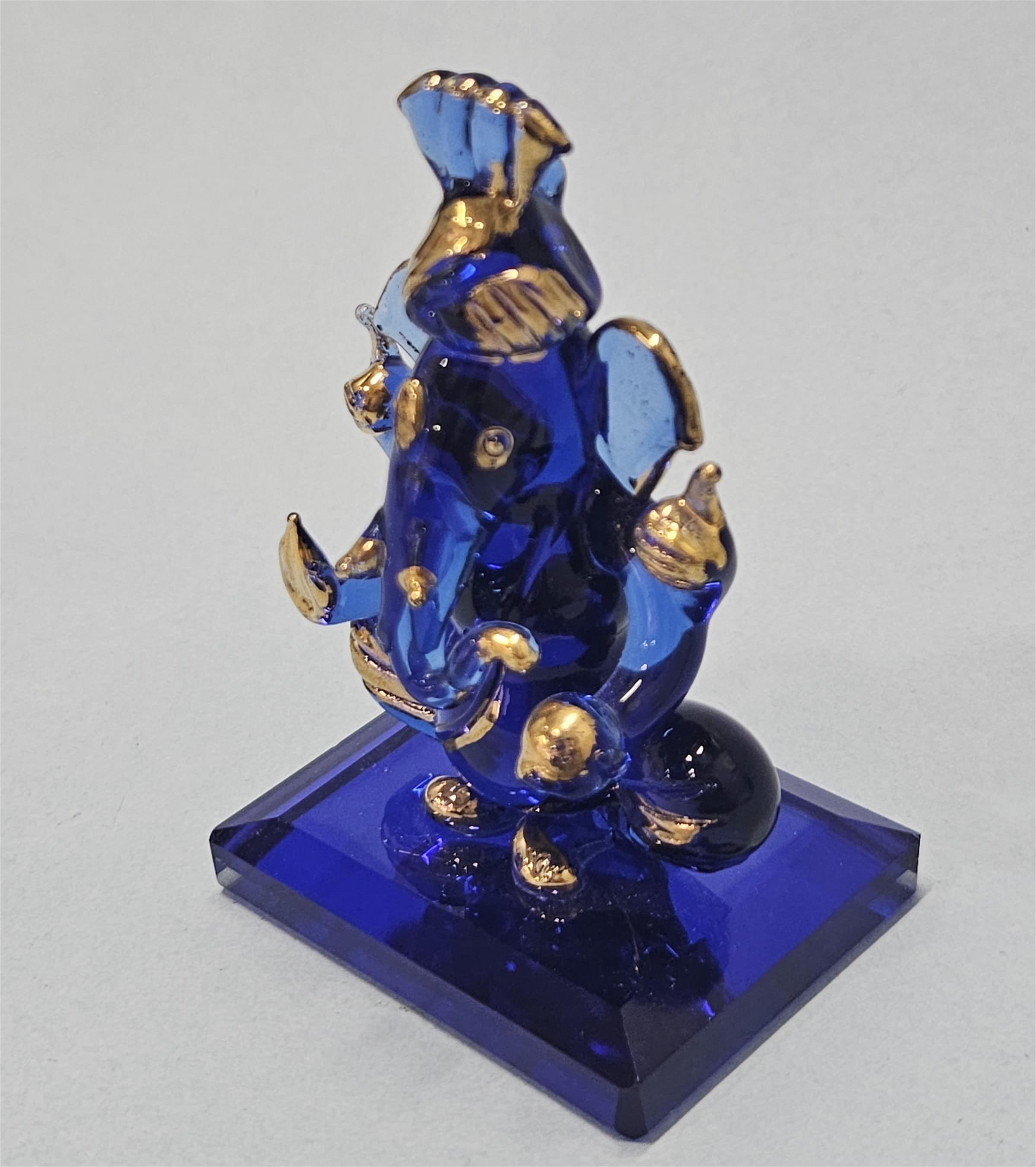 A blue crystal Ganesha idol with gold accents is shown, wearing a pagri. The statue is measured at approximately 2 inches wide and 3 inches tall, and it is displayed against a white background.