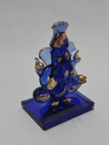 A close-up of a small, intricately designed blue glass Pagri Ganesha idol with gold accents, sitting on a rectangular blue base.