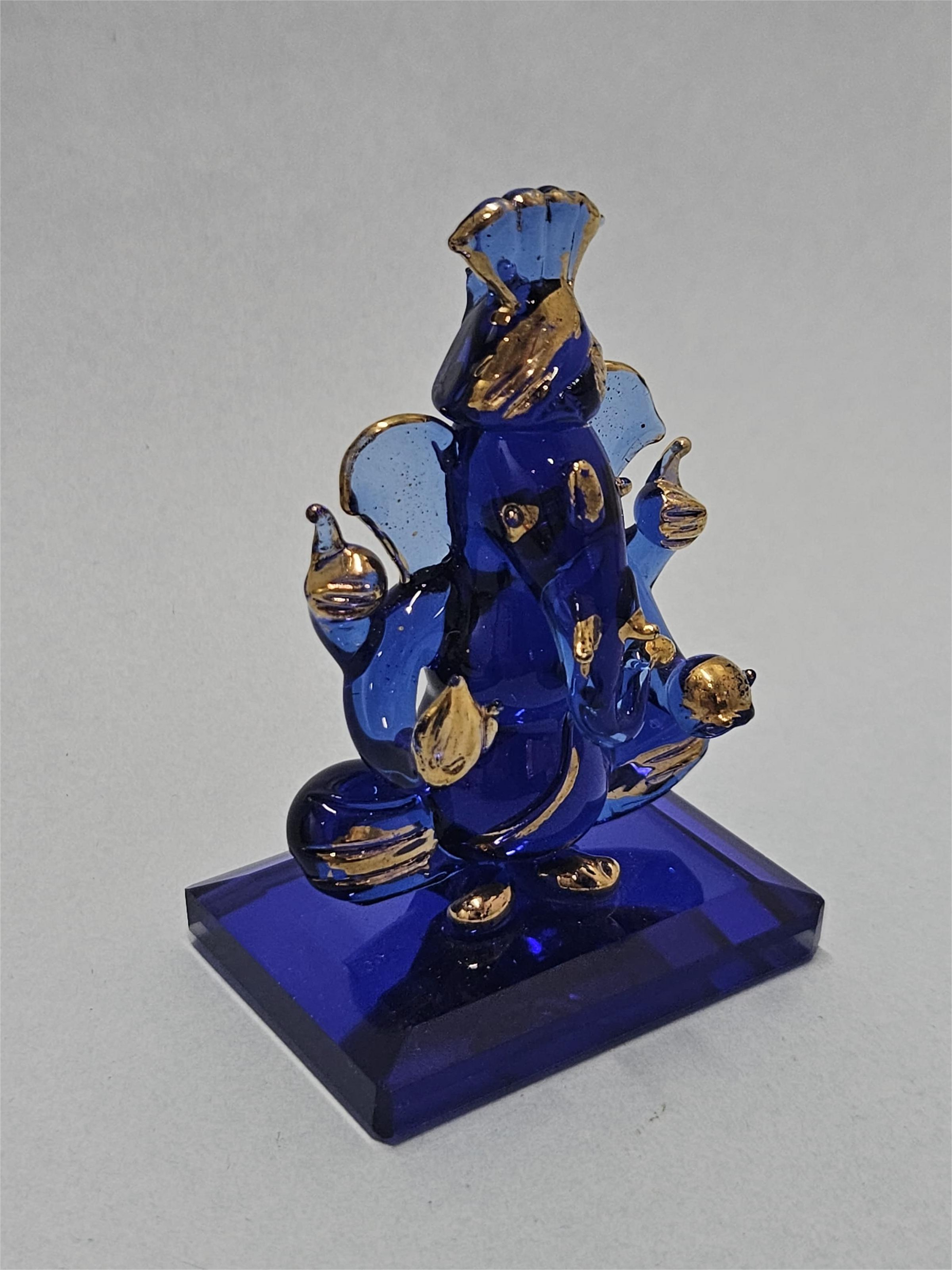 A close-up of a small, intricately designed blue glass Pagri Ganesha idol with gold accents, sitting on a rectangular blue base.