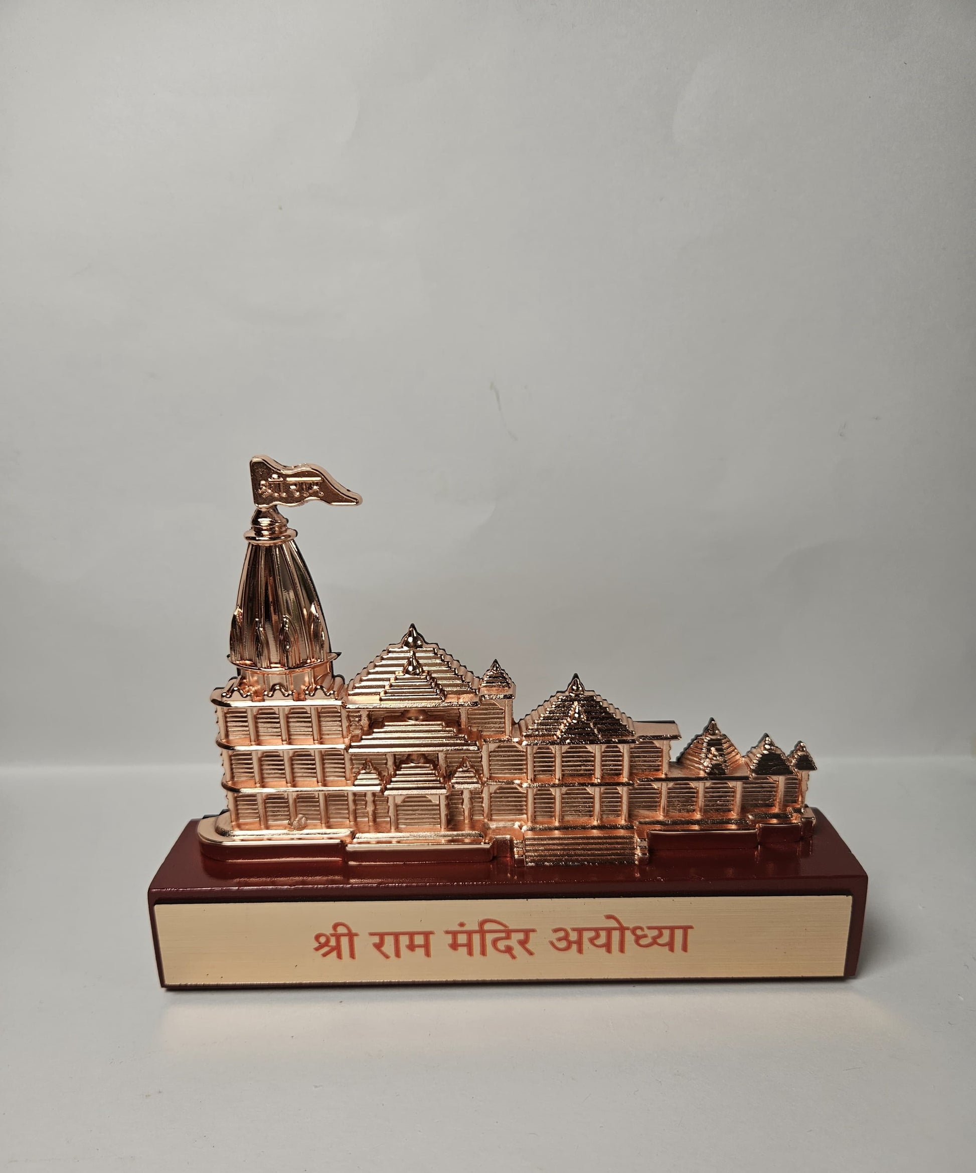 A beautifully sculpted model of the Shri Ram Mandir in Ayodhya, with a reflective copper hue, standing on a base clearly labeled with its identity in Devanagari.