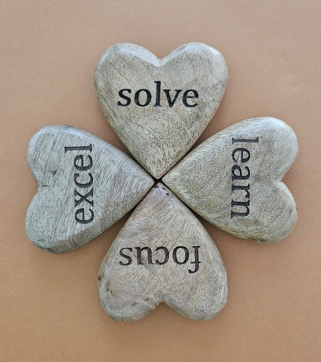 A set of four heart-shaped mango wood tokens with the words 'solve', 'excel', 'learn', and 'focus' engraved on them, arranged in a square on a beige background.