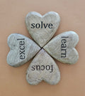 A set of four heart-shaped mango wood tokens with the words 'solve', 'excel', 'learn', and 'focus' engraved on them, arranged in a square on a beige background.
