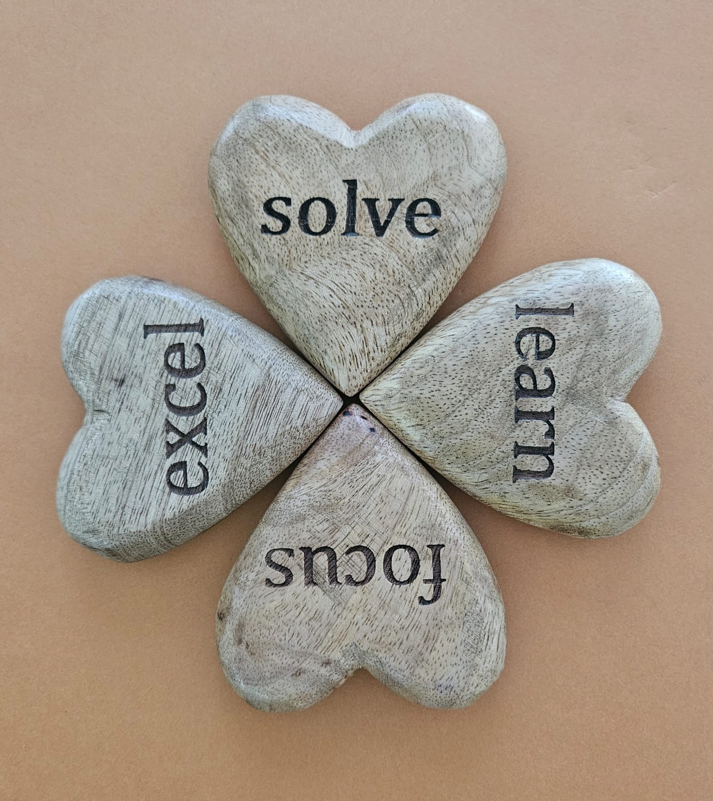 A set of four heart-shaped mango wood tokens with the words 'solve', 'excel', 'learn', and 'focus' engraved on them, arranged in a square on a beige background.
