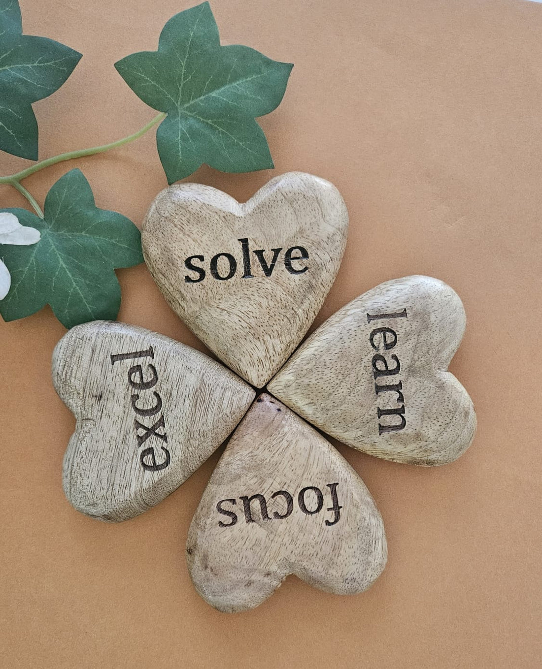 A set of four heart-shaped mango wood tokens with the words 'solve', 'excel', 'learn', and 'focus' engraved on them, arranged in a square on a beige background.