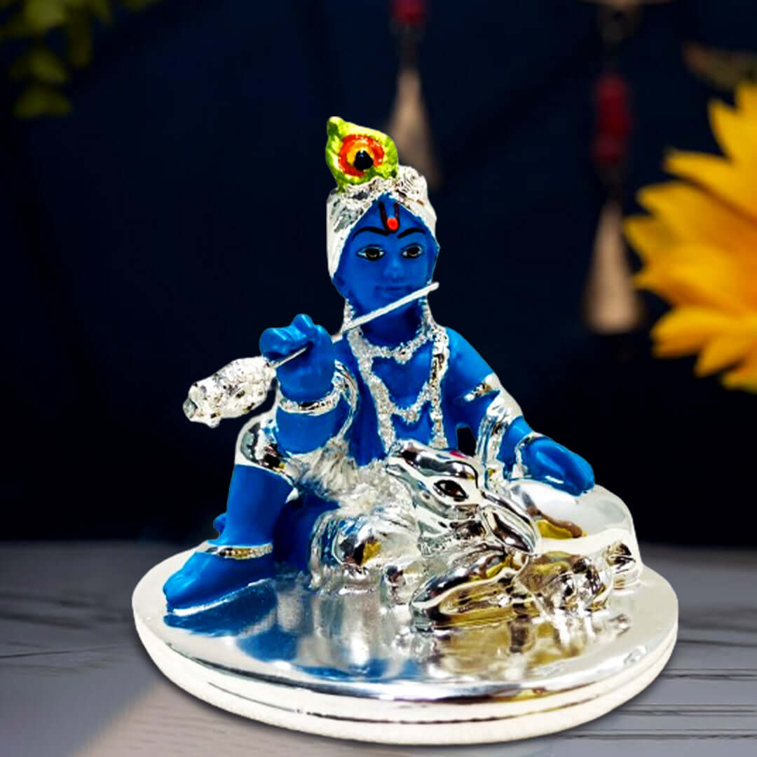 A blue idol of Bal Krishna, adorned with silver ornaments and a peacock feather, sitting and playing a silver flute, with a silver cow resting peacefully beside him, all placed on a silver-colored base.