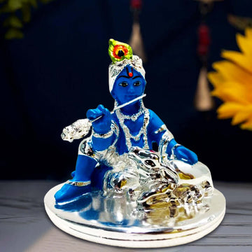 A blue idol of Bal Krishna, adorned with silver ornaments and a peacock feather, sitting and playing a silver flute, with a silver cow resting peacefully beside him, all placed on a silver-colored base.