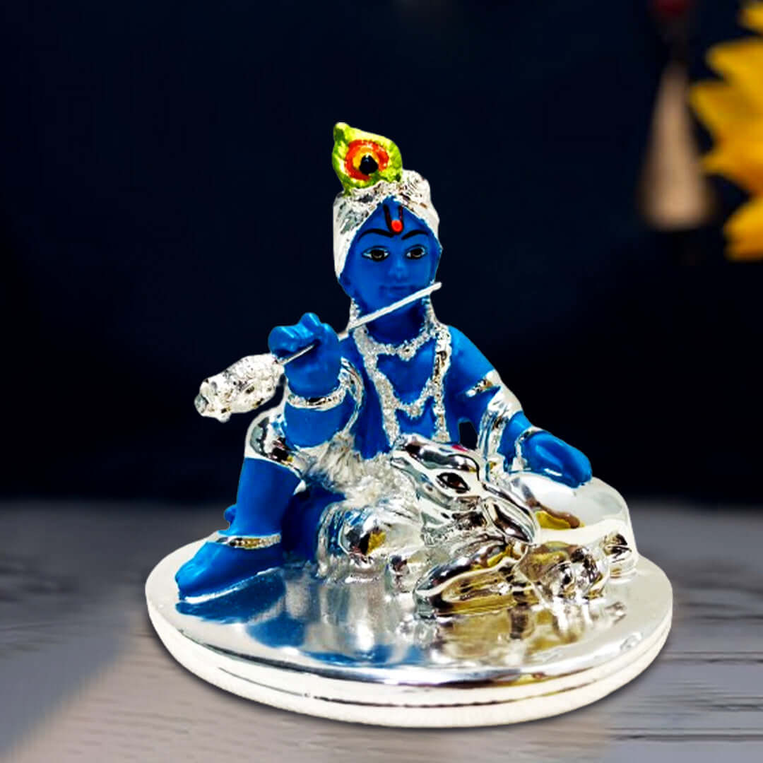 A blue idol of Bal Krishna, adorned with silver ornaments and a peacock feather, sitting and playing a silver flute, with a silver cow resting peacefully beside him, all placed on a silver-colored base.