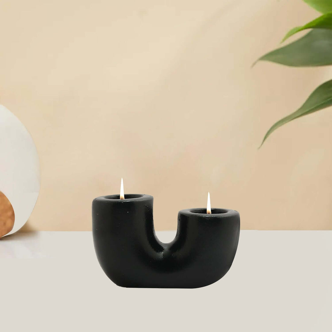 A black U-shaped candle holder with two lit candles placed on it, set against a neutral background with a plant to the side.