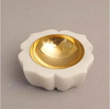 A white, carved stone diya or bowl with a fluted, lotus-like shape, holding a shiny, golden-colored metal cup in the center, all placed on a gray background.