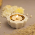 A Petals Marble Diya is a carved white marble candle holder shaped like a lotus flower, with a lit tea light candle in a golden cup at its center. The diya is placed on a gray surface, with blurred white flowers and pearls visible in the background.
