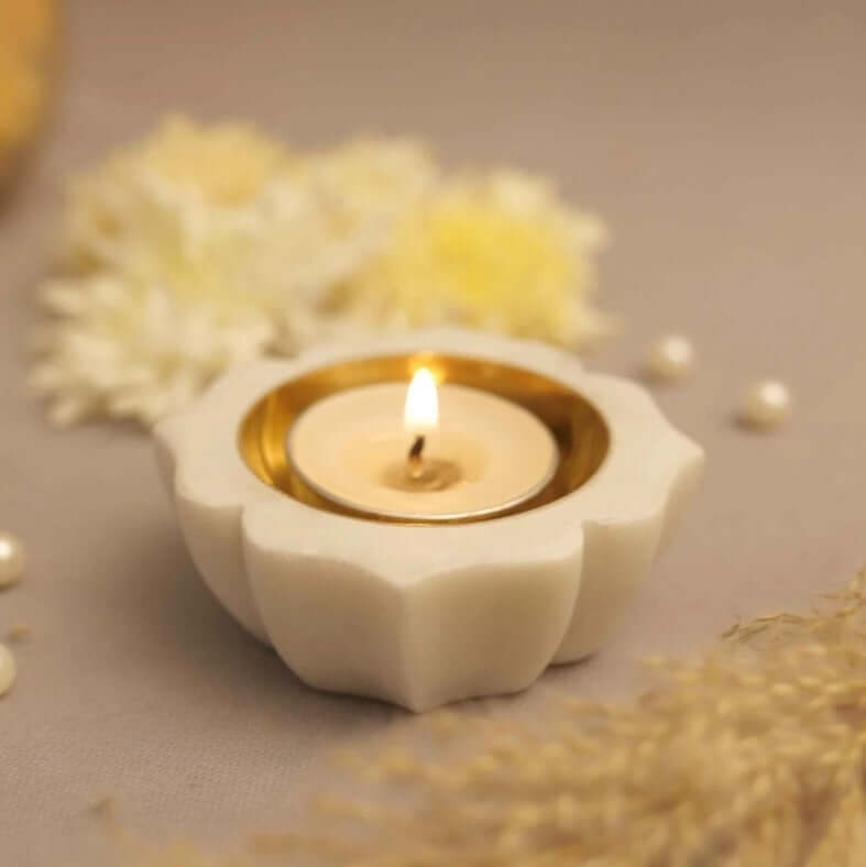 A Petals Marble Diya is a carved white marble candle holder shaped like a lotus flower, with a lit tea light candle in a golden cup at its center. The diya is placed on a gray surface, with blurred white flowers and pearls visible in the background.