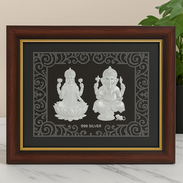 A brown framed silver relief featuring Hindu deities Lakshmi seated on a lotus on the left, and Ganesha seated with his vahana mouse on the right, with '999 SILVER' inscribed below them.