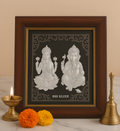 A framed silver-colored relief depicting Hindu deities Lakshmi and Ganesh, seated on lotus flowers with '999 SILVER' inscribed below, displayed alongside a lit diya, a brass bell, and fresh orange and yellow marigold flowers.