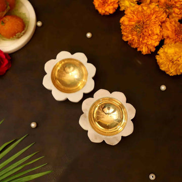 A pair of 8 petals Marble Diyas, featuring a white flower-shaped marble base with a polished golden bowl-like interior. The diyas are placed on a dark surface with scattered white pearls, orange marigold flowers, a green palm leaf, and a white plate of sweets.