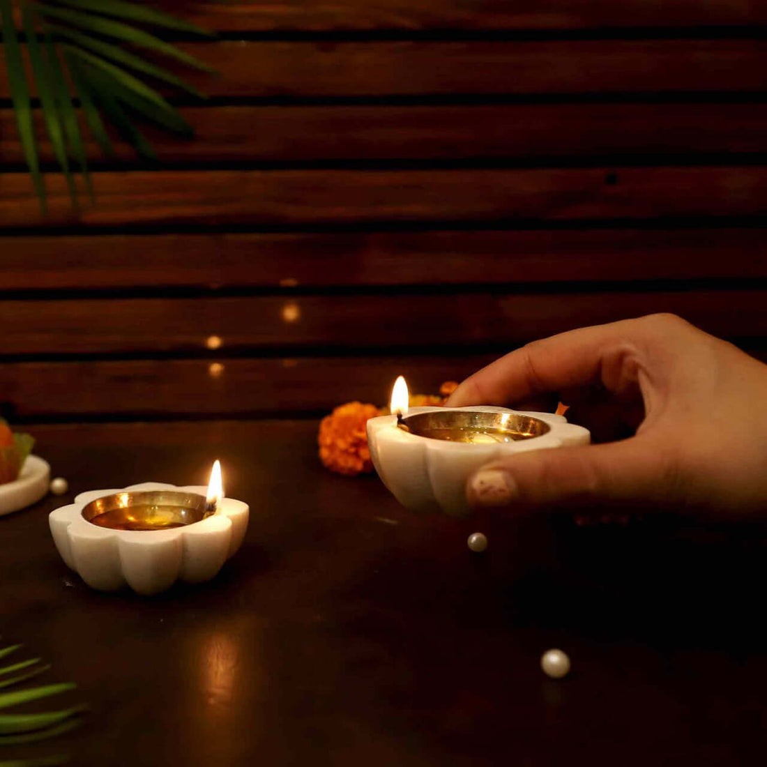 A pair of 8 petals Marble Diyas, featuring a white flower-shaped marble base with a polished golden bowl-like interior. The diyas are placed on a dark surface with scattered white pearls, orange marigold flowers, a green palm leaf, and a white plate of sweets.