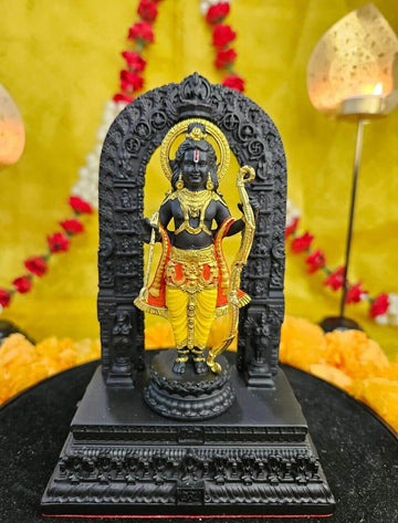 A meticulously crafted, dark-colored idol of Ram Lalla from Ayodhya, adorned with gold ornaments and yellow dhoti, stands under an elaborate black arch on a tiered base, with a golden and red floral background.