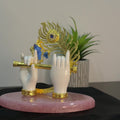 A close-up of a gold-plated Lord Krishna Hands idol featuring two white hands holding a golden flute, with a vibrant peacock feather intricately designed above, all resting on a pinkish-marble base with a green plant in the soft-focus background.