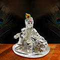 A silver-plated idol of Bal Krishna, a form of Lord Krishna as a child, seated with a silver cow. The idol features intricate details, including a peacock feather on Krishna's head, a flute, and traditional attire, all set on a round base with peacock feather motifs in the background.