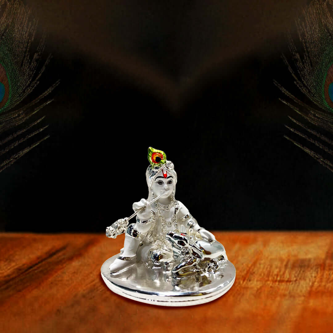 A silver idol of Lord Bal Krishna (Ladoo Gopal) seated and playing a flute, adorned with a colorful peacock feather on his crown, with a small silver cow idol beside him, set on a wooden surface against a dark background.