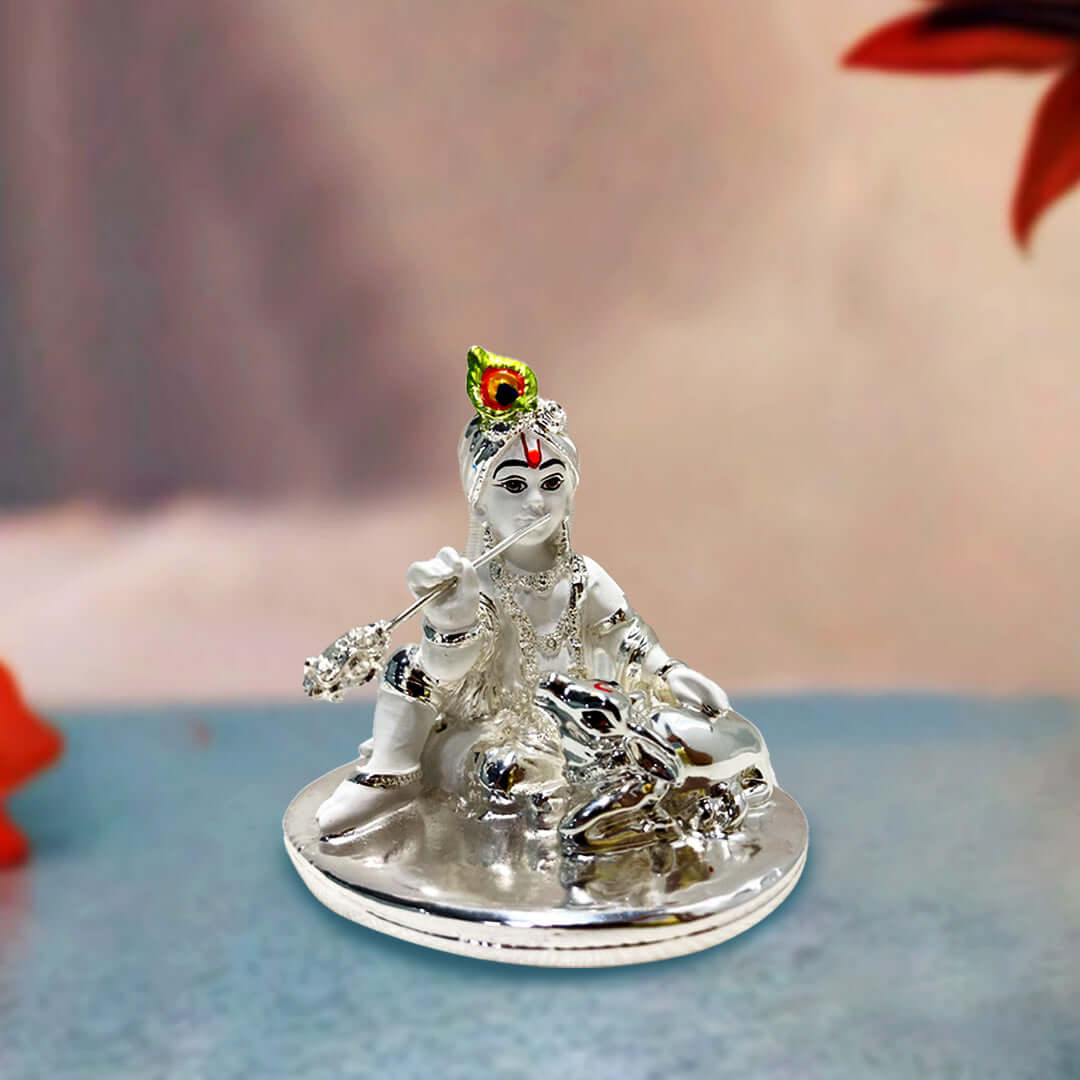 A silver-colored idol of Bala Krishna (child Krishna) sitting, holding a flute, with a small silver cow resting beside him on a polished base. The background is softly blurred.