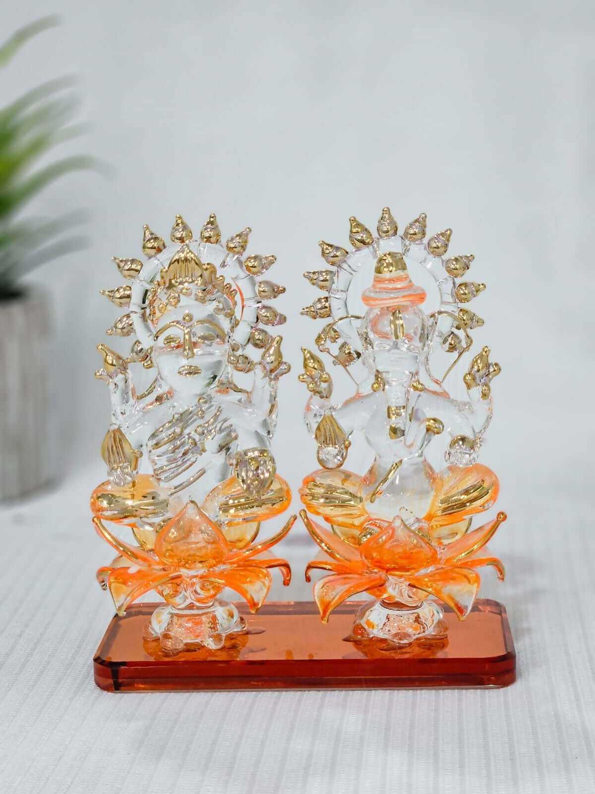 A pair of clear glass statues of the Hindu deities Lakshmi and Ganesha, seated on orange lotus-shaped bases with gold details. Lakshmi is on the left and Ganesha is on the right, both adorned with gold accents on their crowns and attire. The statues are set on a rectangular, reddish-brown base.