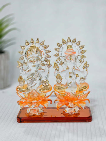 A pair of clear glass statues of the Hindu deities Lakshmi and Ganesha, seated on orange lotus-shaped bases with gold details. Lakshmi is on the left and Ganesha is on the right, both adorned with gold accents on their crowns and attire. The statues are set on a rectangular, reddish-brown base.