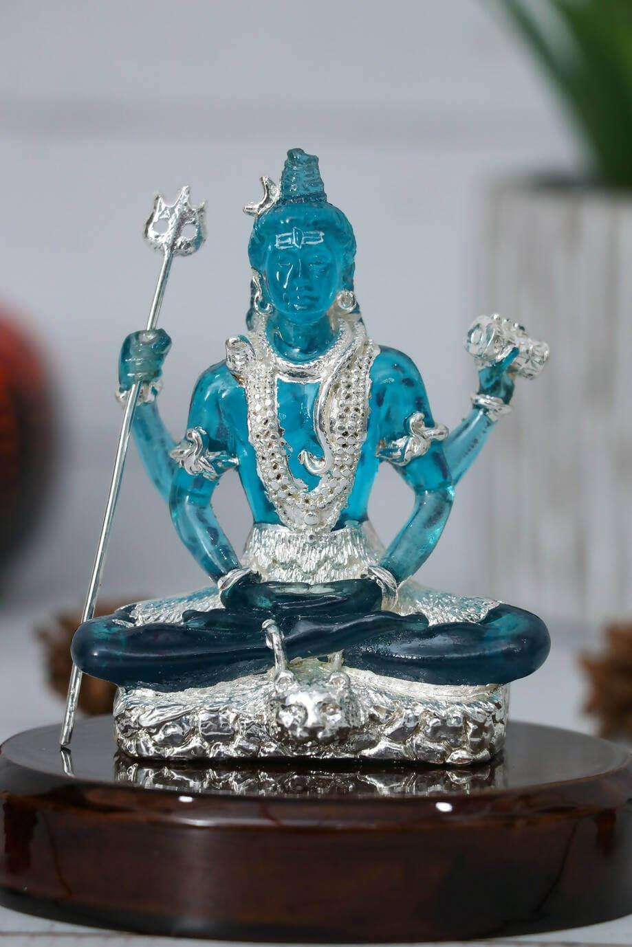 A blue, translucent Lord Shiva idol crafted from crystal or glass, seated in a meditative pose on an ornate silver-colored base, holding a silver trident in one hand and a flower in another, placed on a dark wooden stand.