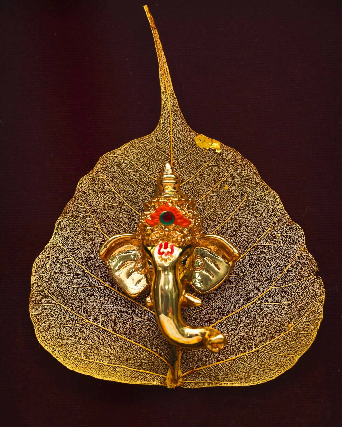 A framed golden Ganesha idol crafted on a delicate, golden leaf, set against a deep burgundy background within an ornate gold frame. The frame rests on a white surface adorned with vibrant yellow marigold flowers and scattered orange petals.